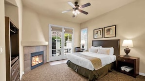 A hotel room with a bed, a fireplace, a ceiling fan, and a view through a glass door. - Desert Serenity C | Studio Legacy Villa (La Quinta)