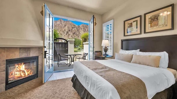 A hotel room with a fireplace, a bed, a balcony with a table and chairs, and framed artwork on the wall. - Desert Serenity C | Studio Legacy Villa (La Quinta)