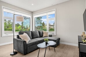 A modern living room with a grey sofa, a small round table, and a TV stand. - 2 King Bed | Sofa Bed | Garage | Min to UofC | NEW (Calgary)