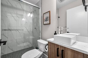 A modern bathroom with a glass shower enclosure, a toilet, a sink with a mirror, and a framed picture on the wall. - King Bed | Min to UofC | NEW (Calgary)