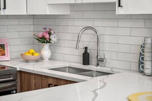A kitchen with white tiles, a stainless steel sink, a vase with flowers, and a bowl of fruit. - King Bed | Min to UofC | NEW (Calgary)