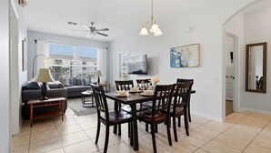 A dining area with a table set for four, a sofa, a ceiling fan, and a TV mounted on the wall. - NEW! Sunshine Stay at Windsor Palms - Near Disney (Kissimmee)