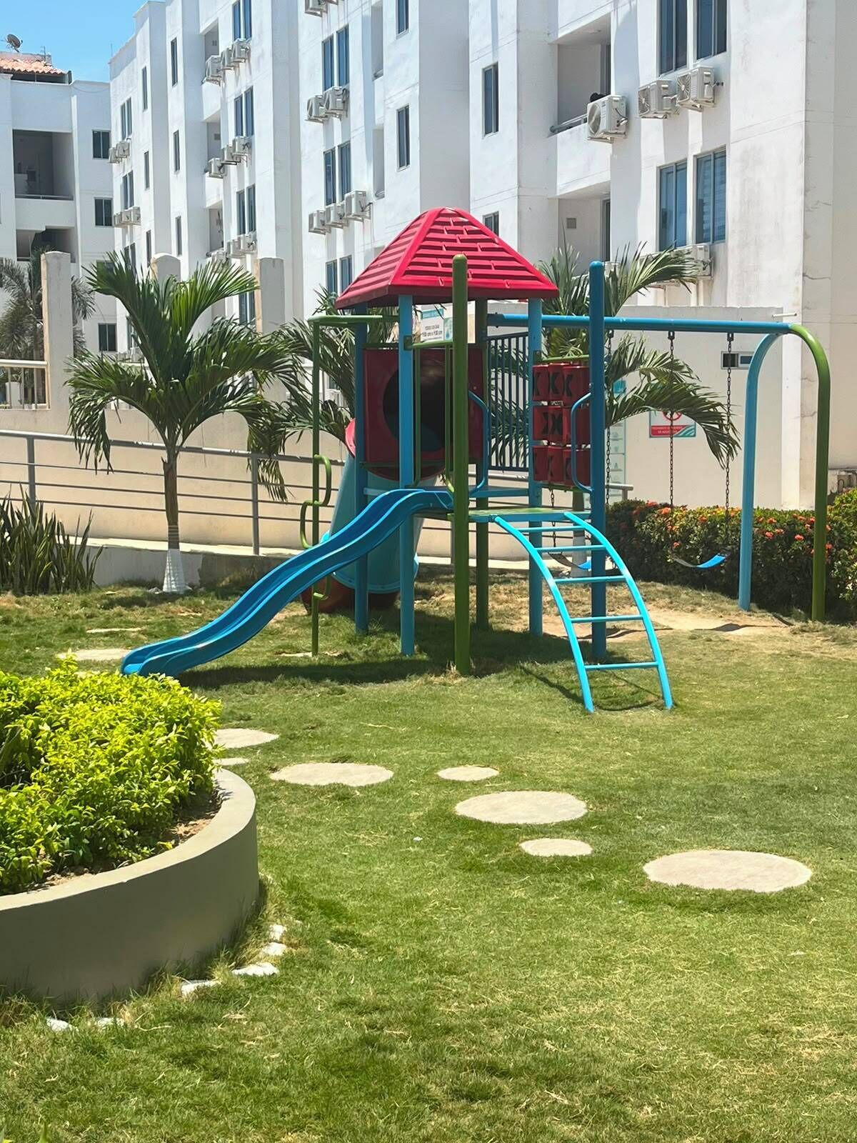 A playground with a red roof and blue slides, surrounded by grass and palm trees, with a building in the background.