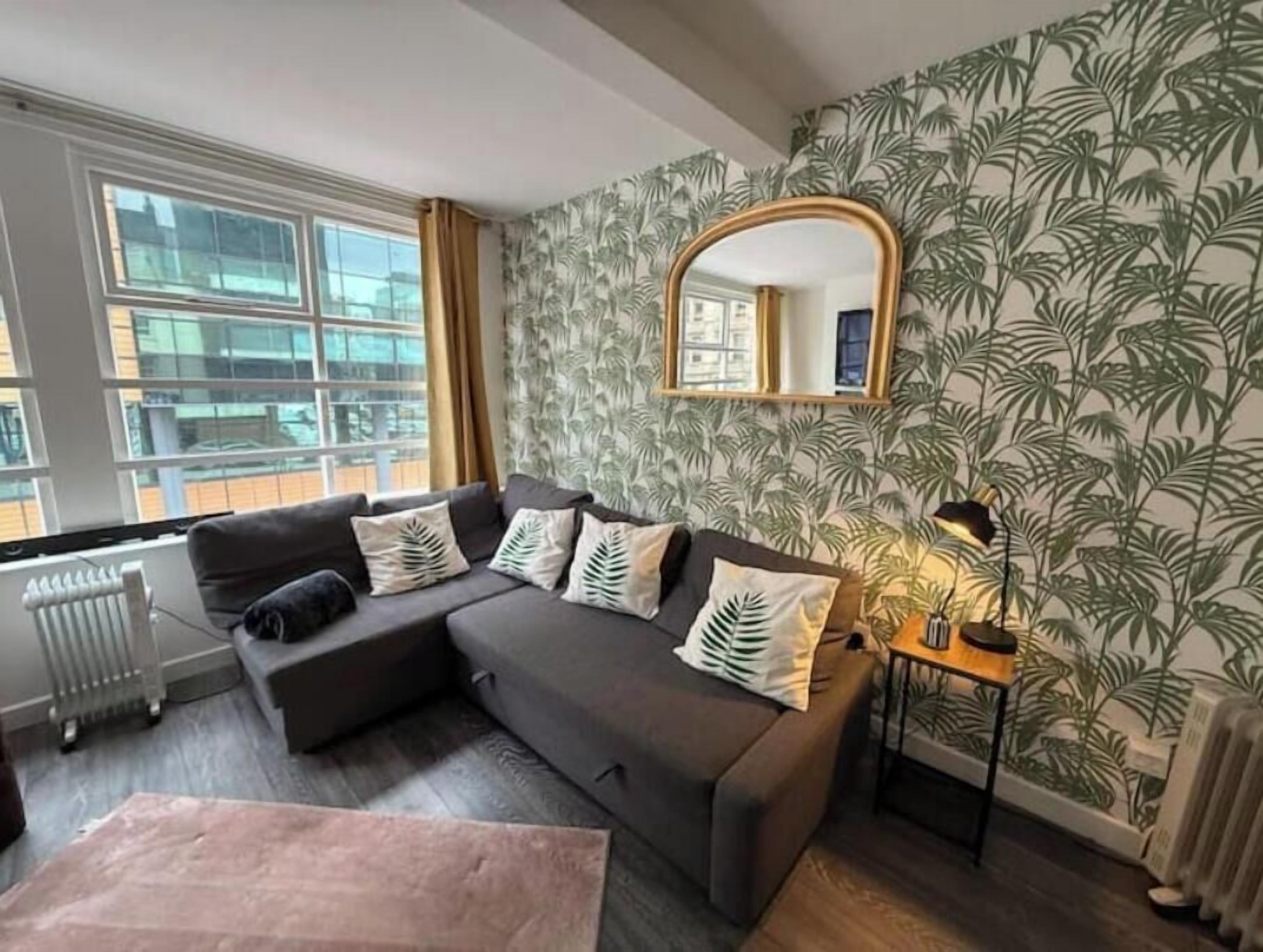 Ein Wohnzimmer mit grauem Sofa, einem Spiegel, einer Heizung und einem Fenster mit Blick auf Gebäude.