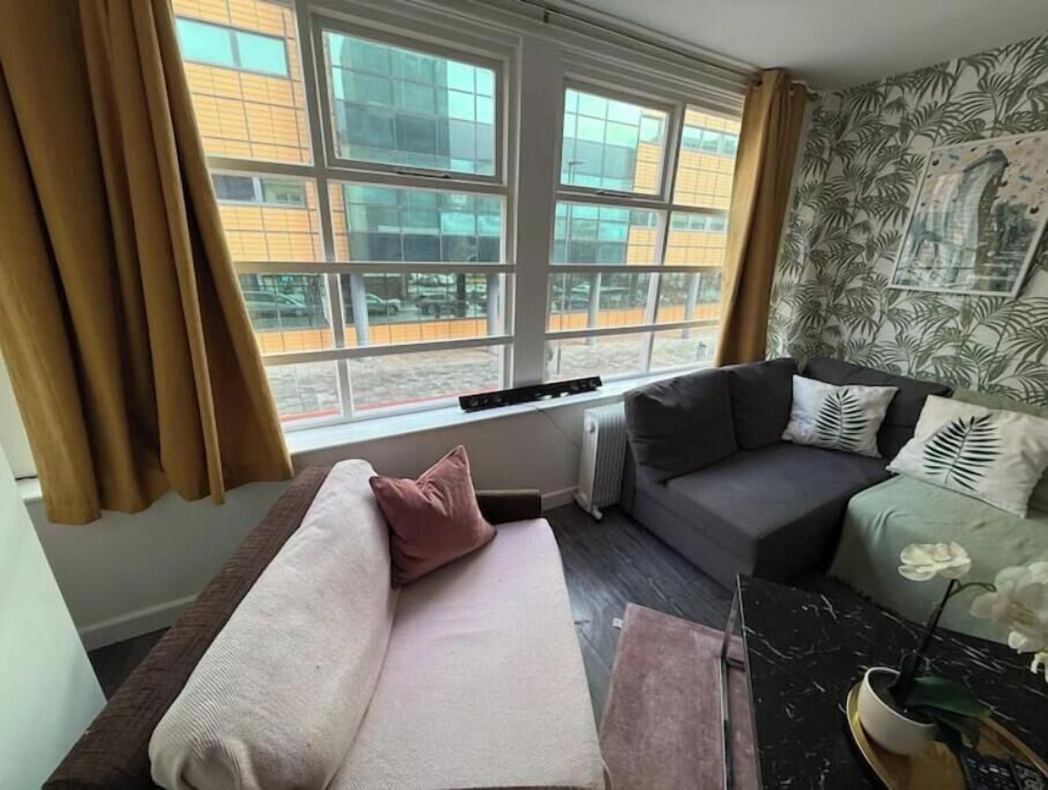 Ein Zimmer mit einer Couch, einem Fenster mit Vorhängen, einem Heizkörper und einer Wand mit einem Blattmuster.