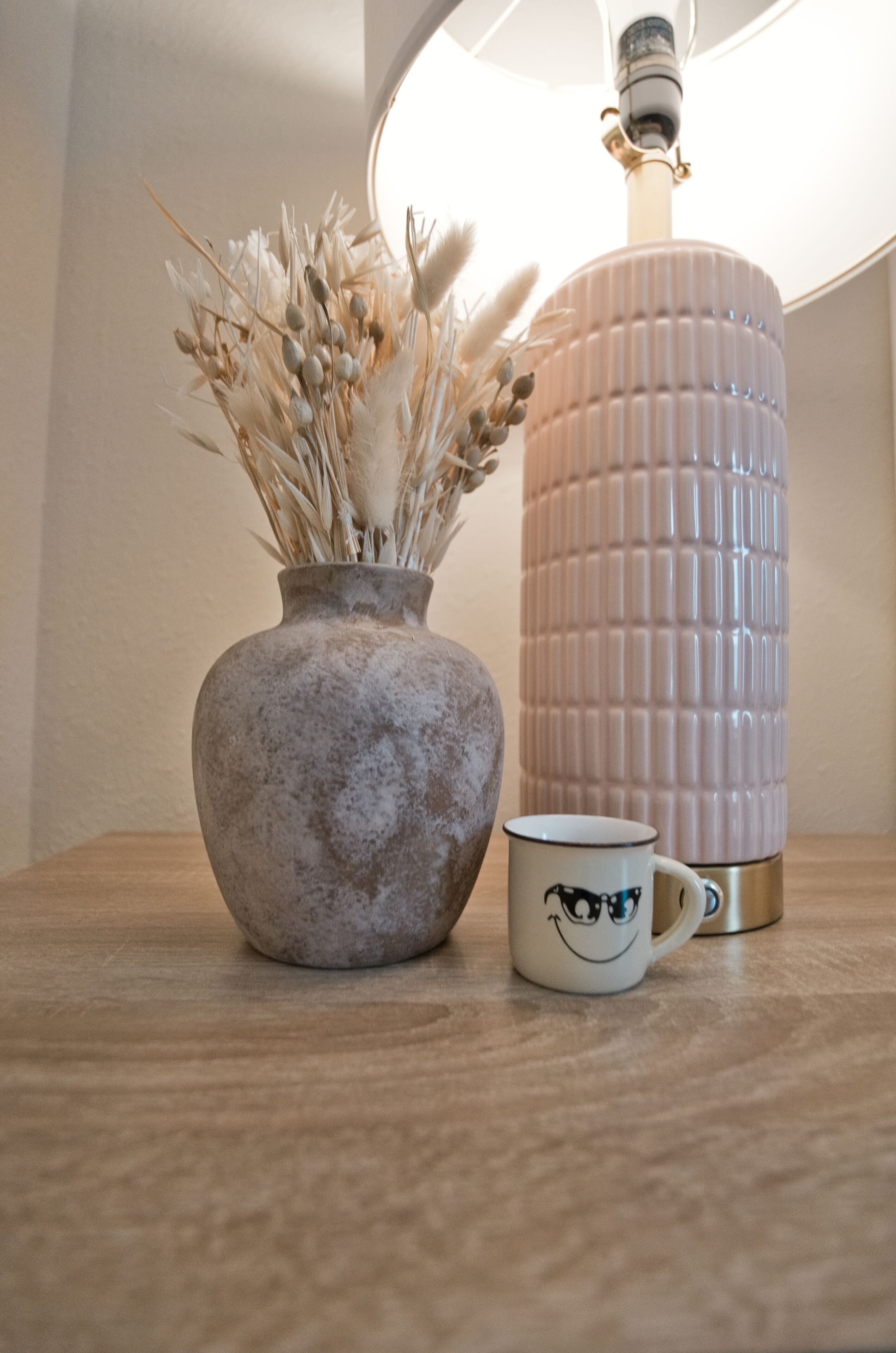 Un florero con plantas secas, una taza y un recipiente cilíndrico sobre una superficie de madera.