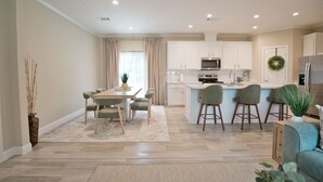 Uma cozinha moderna com armários brancos, uma ilha central com banquetas, uma área de refeições com mesa e cadeiras, e uma sala de estar com sofá e uma planta.
