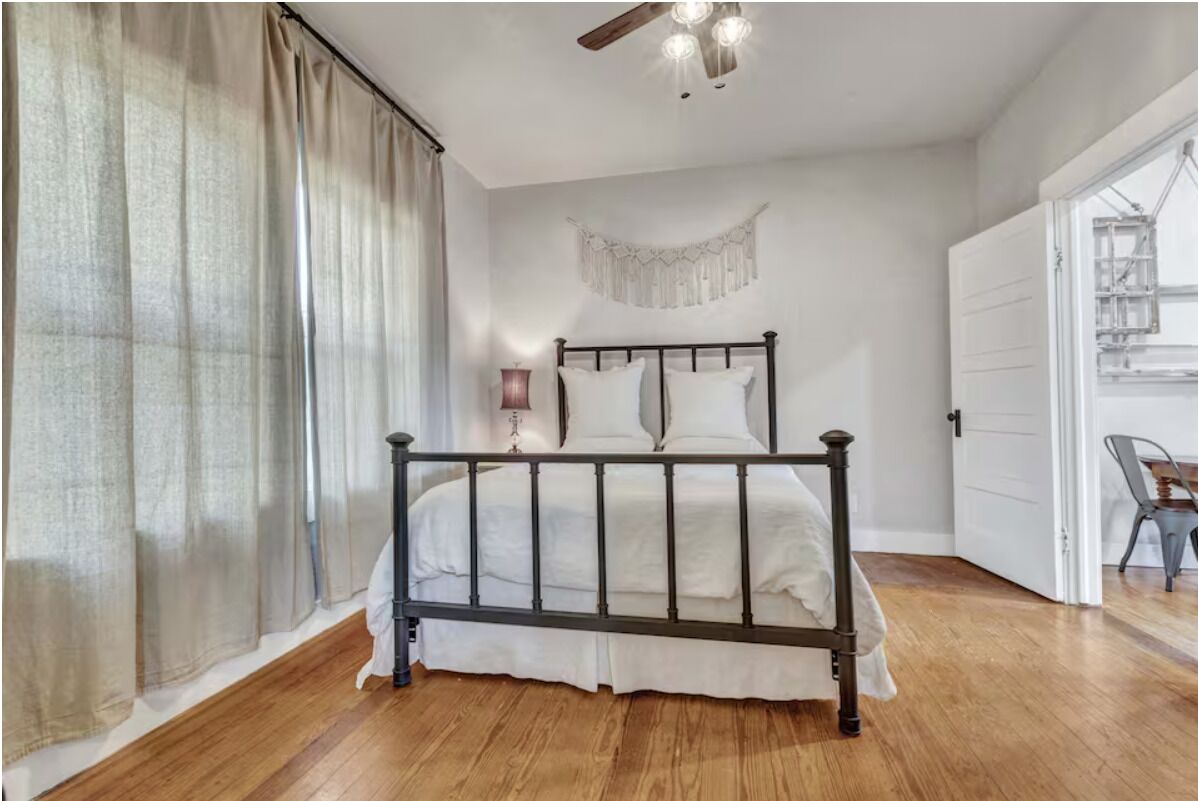 Un dormitorio con cama, cortinas, un macramé colgado en la pared, un ventilador de techo y una puerta que da a otra habitación.
