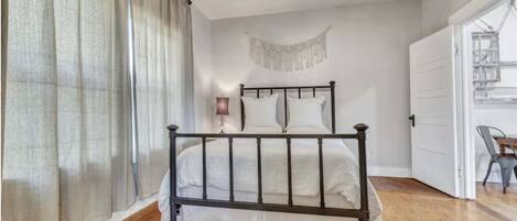 Un dormitorio con cama, cortinas, un macramé colgado en la pared, un ventilador de techo y una puerta que da a otra habitación.
