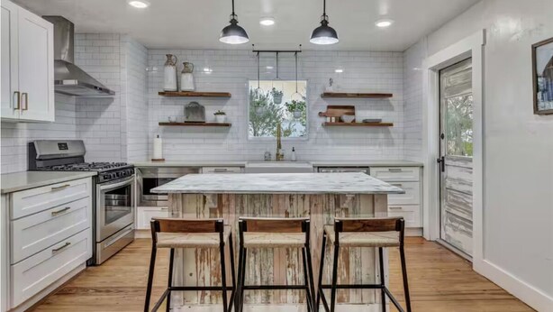 Una cucina moderna con mobili bianchi, un'isola centrale con sgabelli da bar, illuminazione a sospensione e vista dalla finestra.