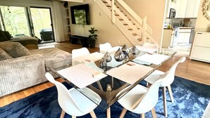 Ein moderner Wohnraum mit Essbereich, der einen Tisch mit Glasplatte und weiße Stühle, eine Treppe und eine Küche im Hintergrund umfasst.