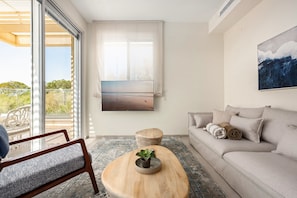 A modern living room with a sofa, a wooden coffee table with a plant, a TV mounted on the wall, and a large window with a view.