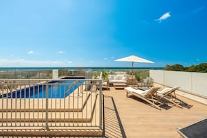A rooftop pool area with a view of the ocean, lounge chairs, and a sun umbrella.