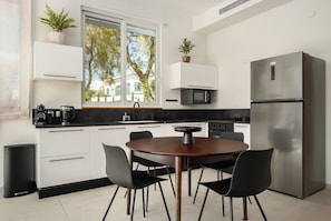 A modern kitchen with white cabinetry, a stainless steel refrigerator, a round wooden dining table with black chairs, and a window with a view of trees.