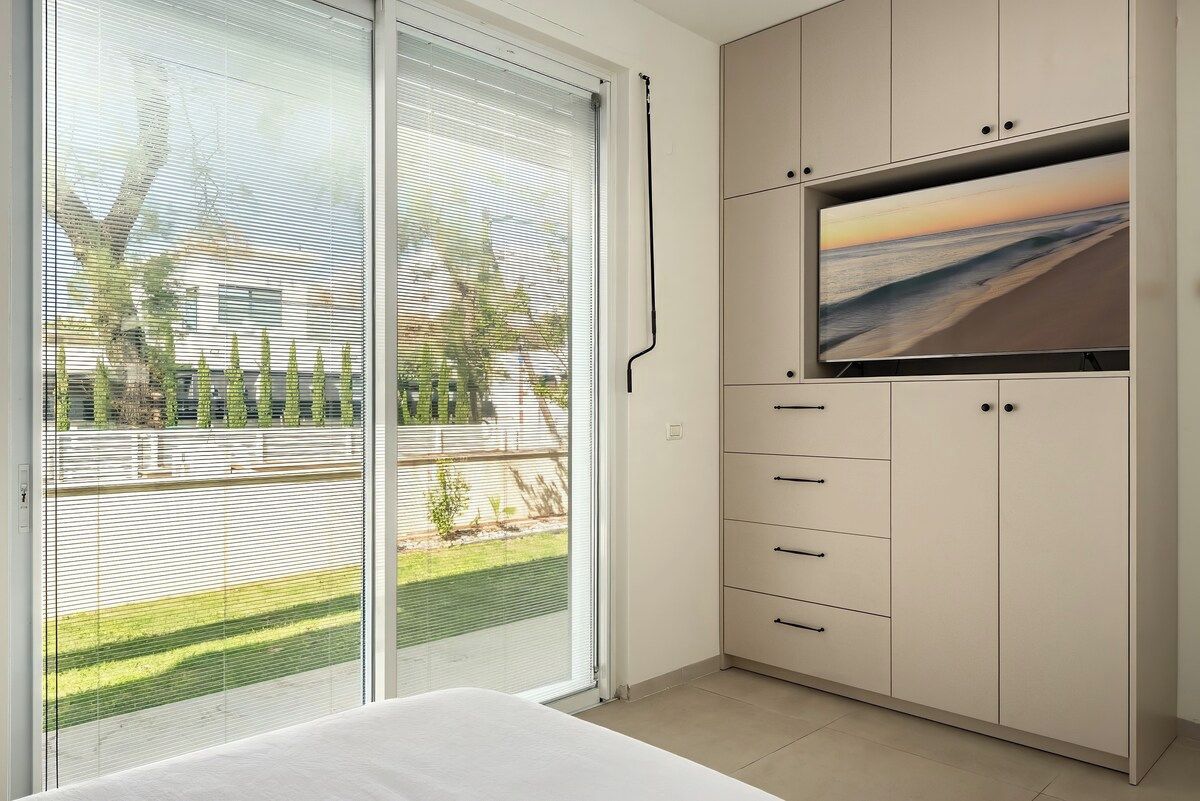Ein Schlafzimmer mit einer Schiebeglastür, die zu einem Balkon führt, einem Bett und einem an einer Kommode befestigten Fernseher.
