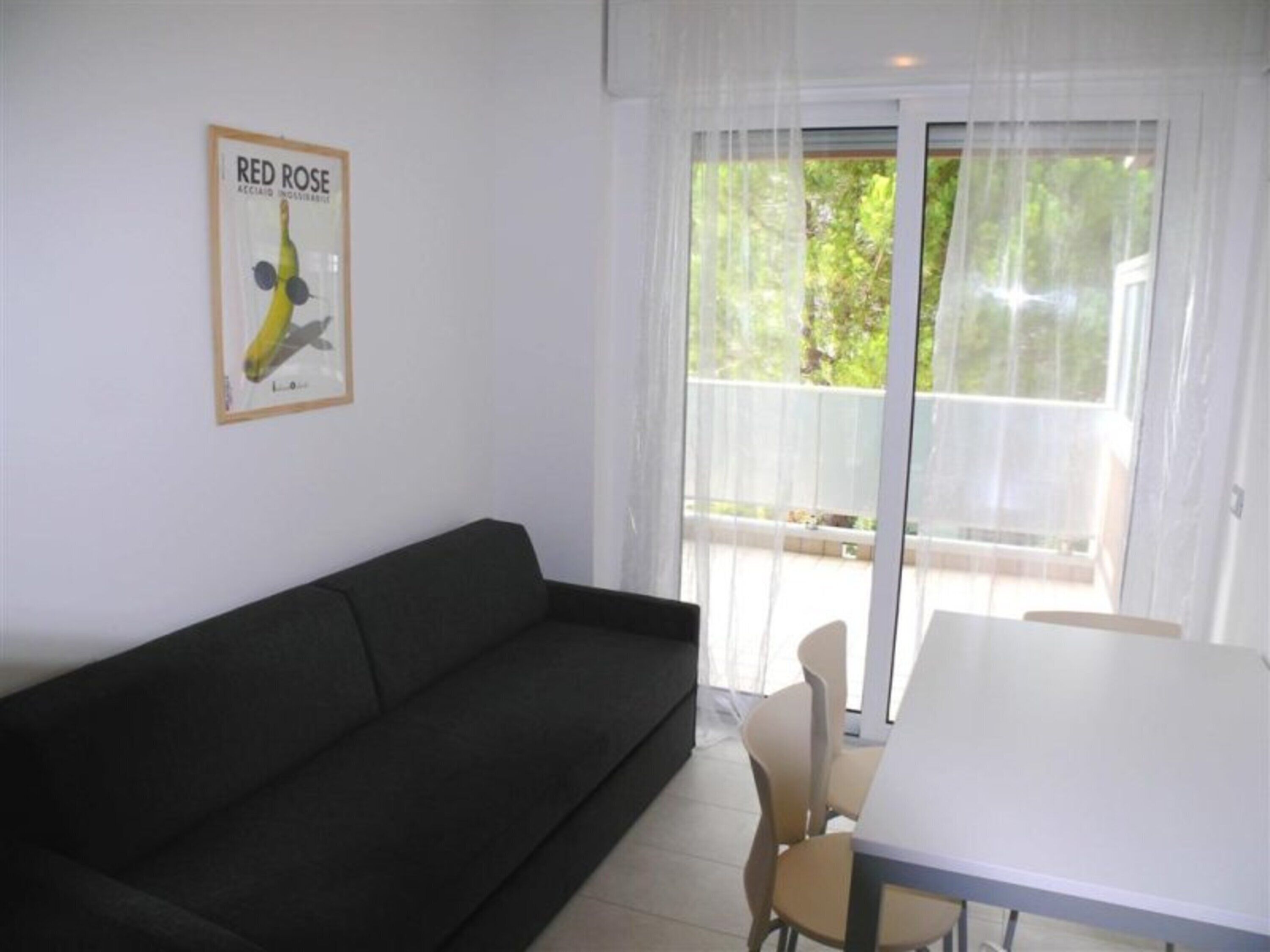 Una habitación con un sofá oscuro, una mesa blanca y una ventana con cortinas translúcidas.