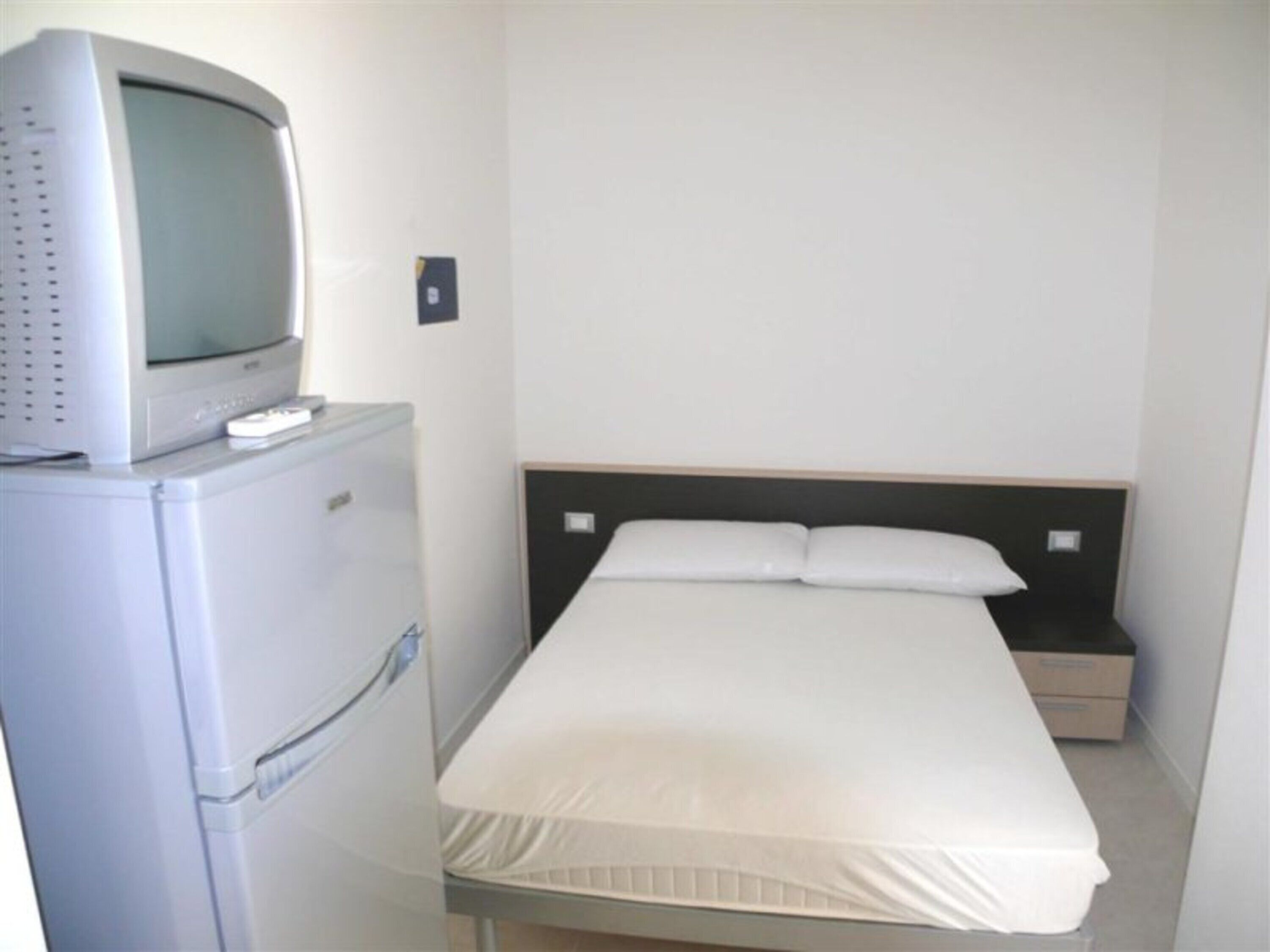 Une petite chambre avec un lit simple, une table de chevet, un téléviseur posé sur un meuble et un réfrigérateur.