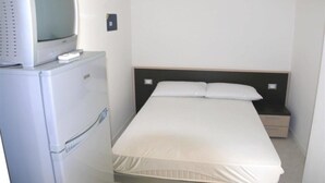 Une petite chambre avec un lit simple, une table de chevet, un téléviseur posé sur un meuble et un réfrigérateur.
