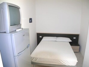 Ein kleines Zimmer mit einem Einzelbett, einem Nachttisch, einem Fernseher auf einem Standfuß und einem Kühlschrank.