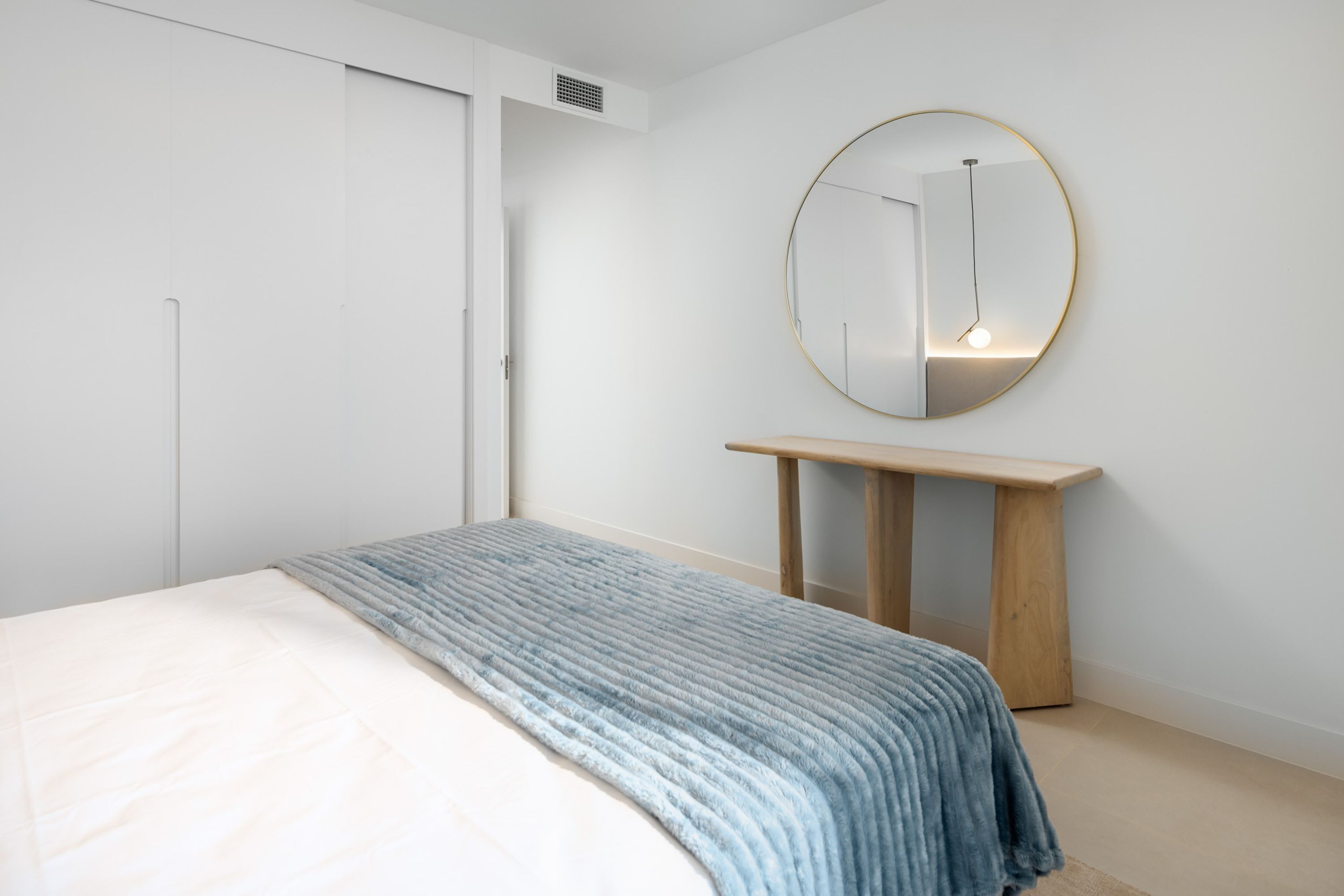 A bedroom with a bed, a round mirror, a wooden console table, and a white wardrobe.