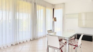 A modern dining area with a glass table and white chairs, large windows with sheer curtains, and a tiled floor. - S362 – Sirolo, wonderful four-room apartment with garden (Sirolo)