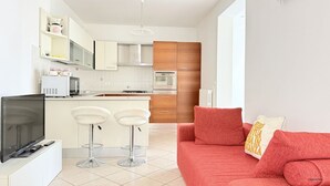 A modern kitchen with white cabinets, a microwave, and a TV on a stand. There is a red sofa with cushions and a small table with two bar stools. - S362 – Sirolo, wonderful four-room apartment with garden (Sirolo)