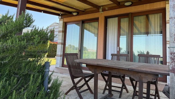 An outdoor patio with a wooden table and chairs, large windows, and a covered area with wooden beams. - S362 – Sirolo, wonderful four-room apartment with garden (Sirolo)