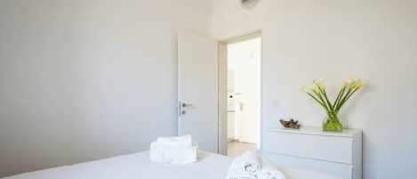 Quarto de hotel com uma cama, mesa de cabeceira com um vaso de flores e porta que dá para outro cômodo.