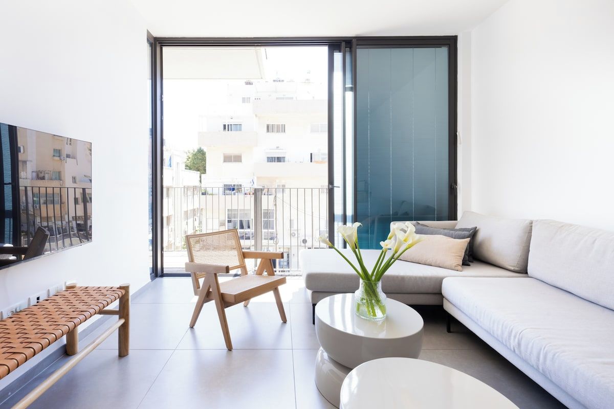Un salon moderne avec un canapé, une table basse, un banc et un fauteuil. Une grande fenêtre offre une vue sur les bâtiments à l’extérieur.
