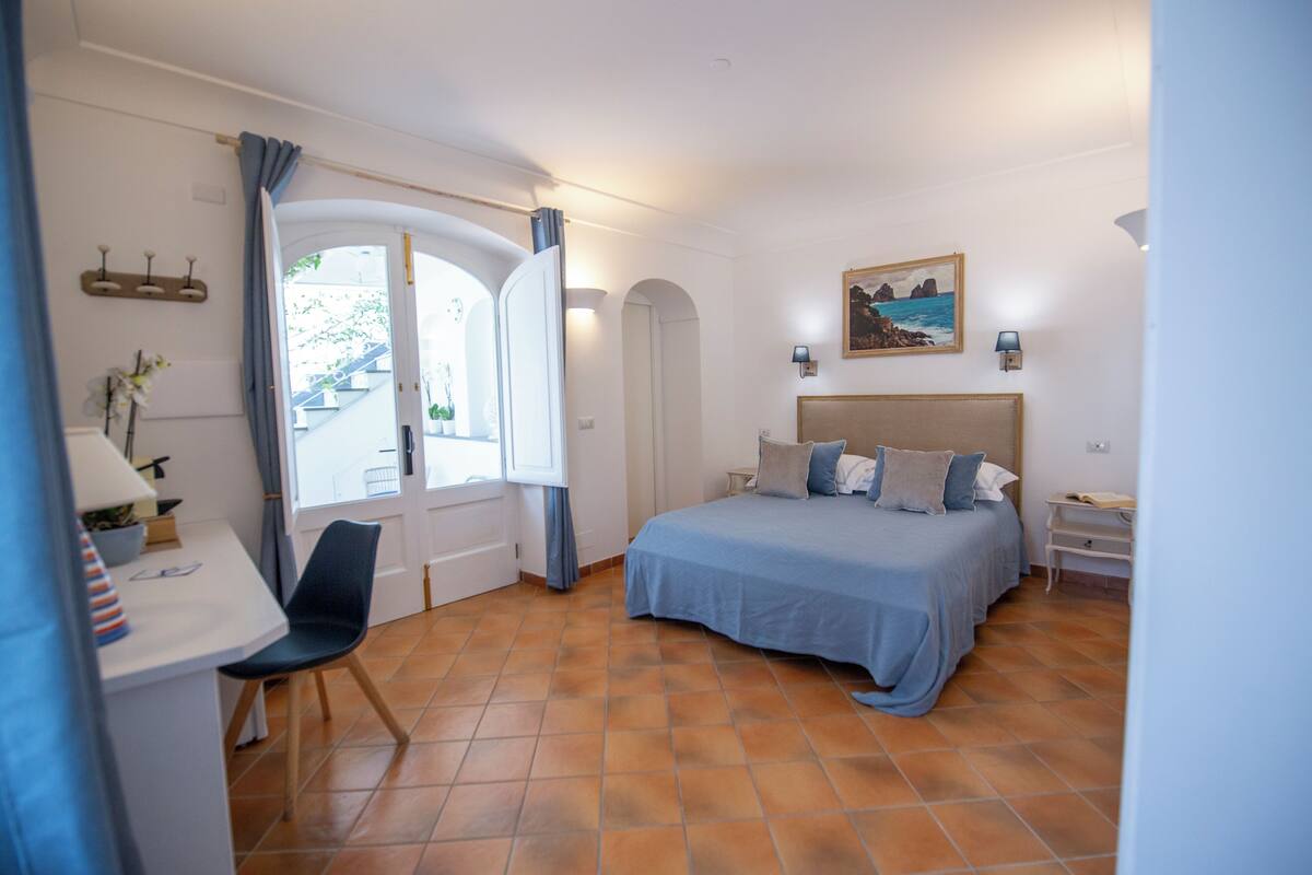 Un dormitorio con cama, escritorio y silla, ventana con cortinas, un cuadro en la pared y piso de baldosas.