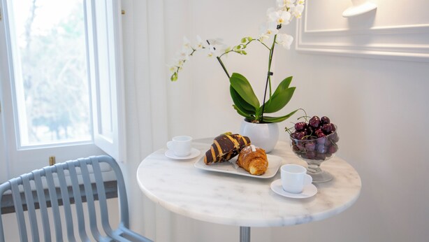 Uma pequena mesa redonda com uma orquídea branca, uma tigela de cerejas, um croissant e uma xícara de café.