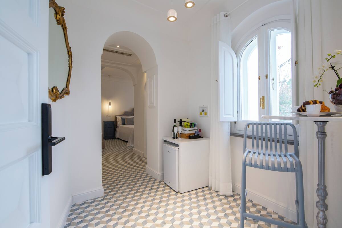 Un pasillo con piso a cuadros, un refrigerador, una silla y una ventana con cortinas.