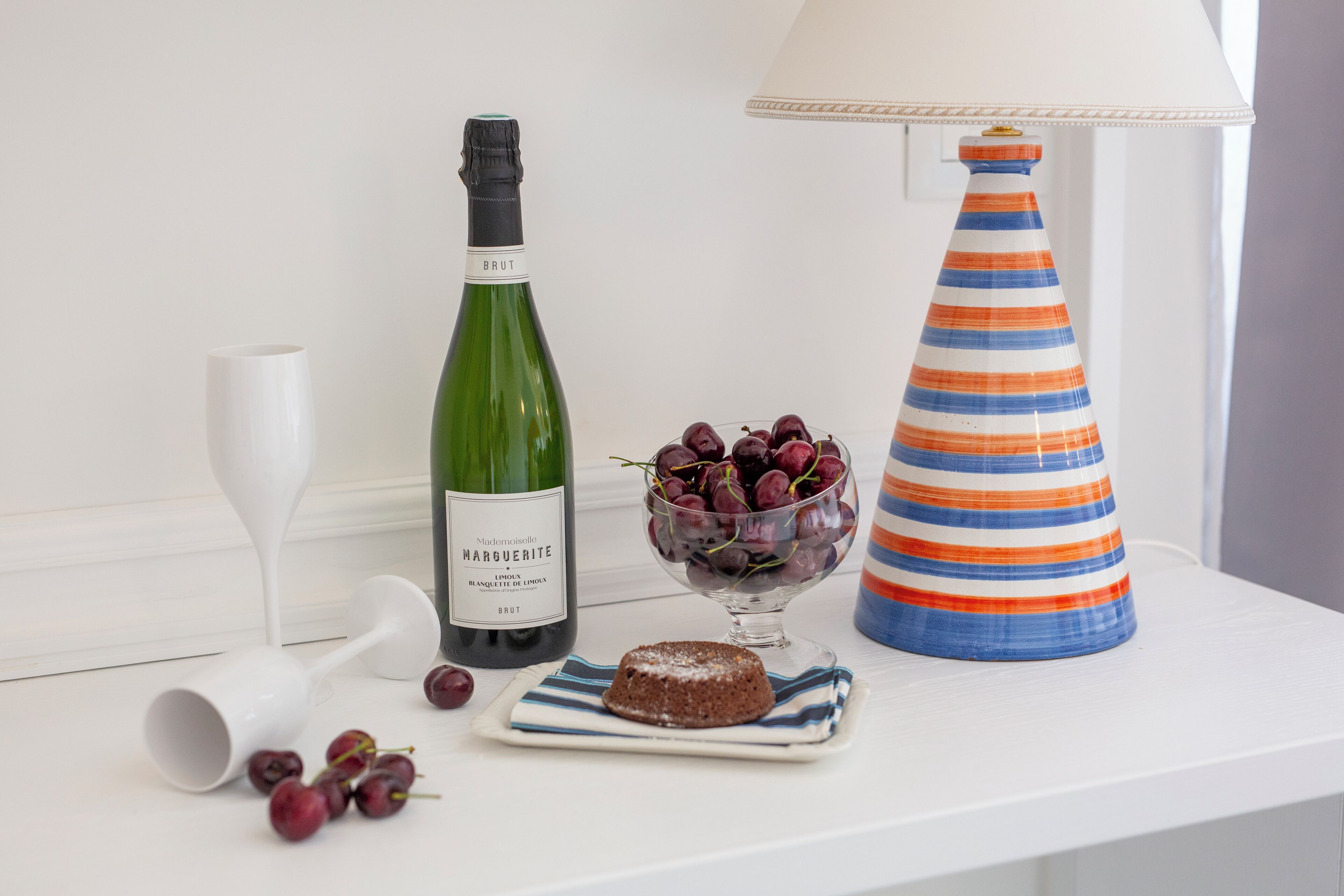 Una mesa con una botella de vino, un cuenco de uvas, una lámpara a rayas y un postre sobre un mantel blanco.