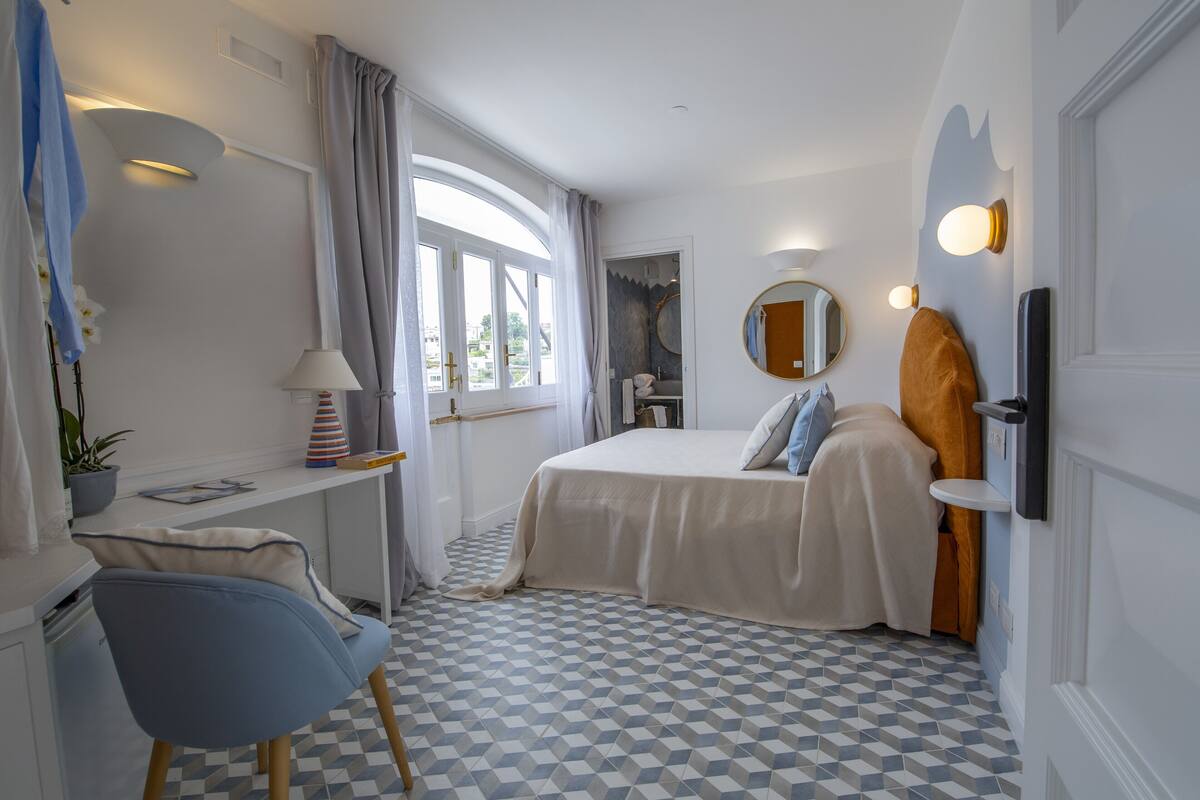 Habitación de hotel con cama, escritorio, silla, ventana con cortinas y piso a cuadros.