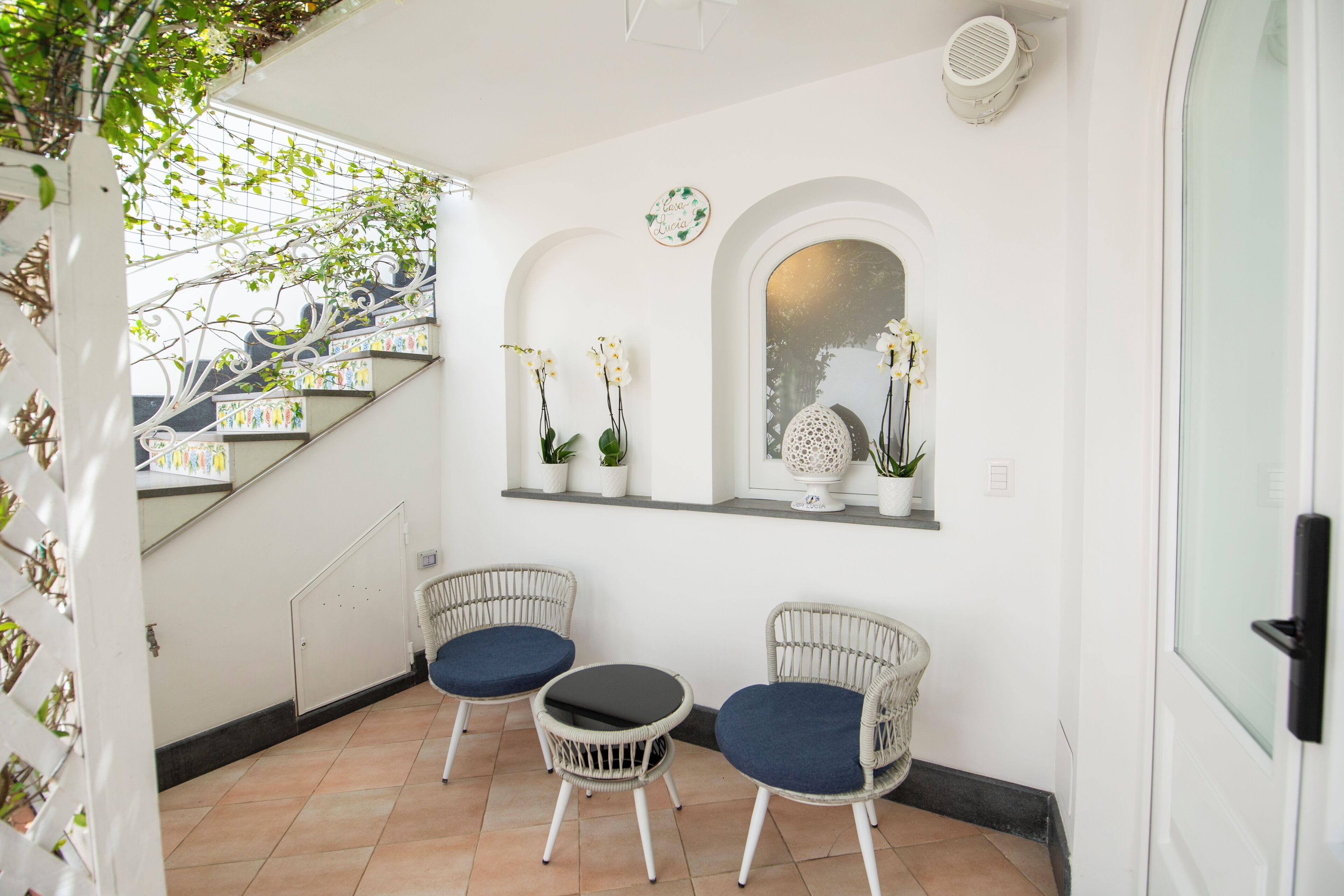 Ein Zimmer mit einer Treppe, zwei Korbstühle mit blauen Kissen, einem kleinen Tisch und einer Wand mit Topfpflanzen und einem Spiegel.