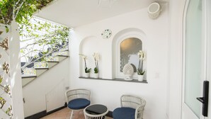 Quarto com escada, duas cadeiras de vime com almofadas azuis, uma mesa pequena e uma parede decorativa com plantas em vasos e um espelho.