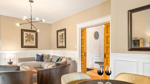 Una sala de estar con sofá, sillones, una mesa de centro, una araña, obras de arte enmarcadas, una mesa de comedor con copas de vino y un espejo.