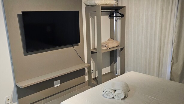 A modern hotel room with a bed, a flat-screen TV mounted on the wall, a built-in air conditioning unit, and a window with curtains. - Flat 109 Norte Novo e Completo 204 (Brasília)