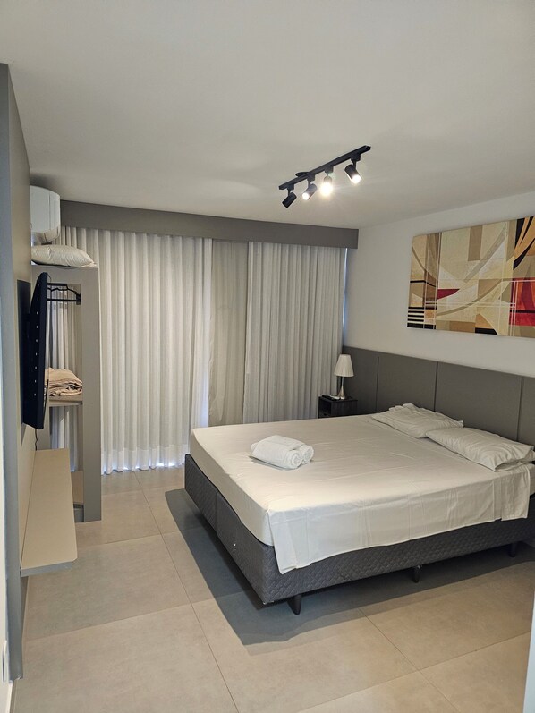 A modern bedroom with a large bed, a flat-screen TV mounted on the wall, a bedside table, and a window with curtains. - Flat 109 Norte Novo e Completo 204 (Brasília)