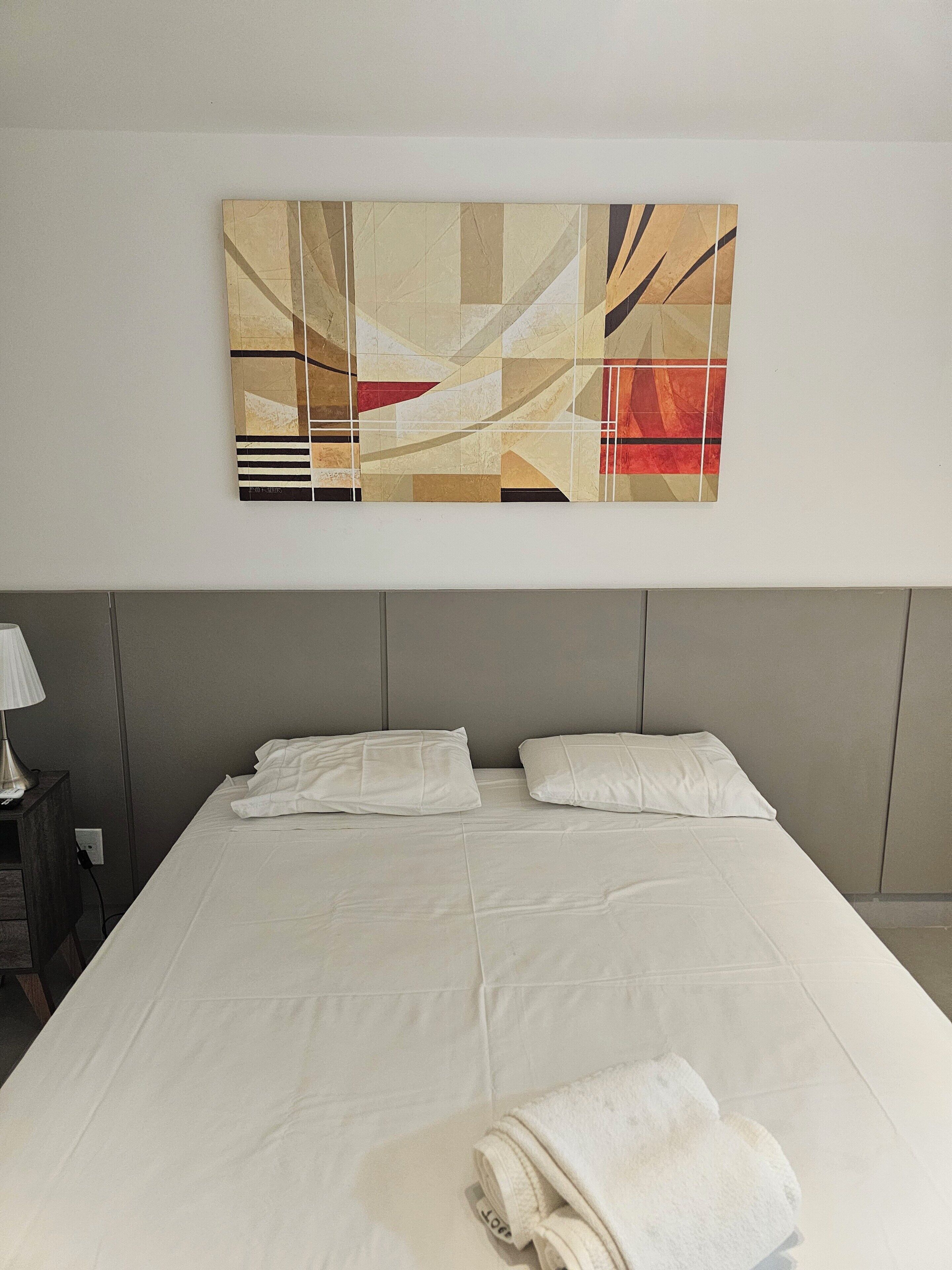 Hotelkamer met een groot bed, twee kussens, een opgevouwen handdoek, een nachtkastje met een lamp, en een abstract schilderij aan de muur.