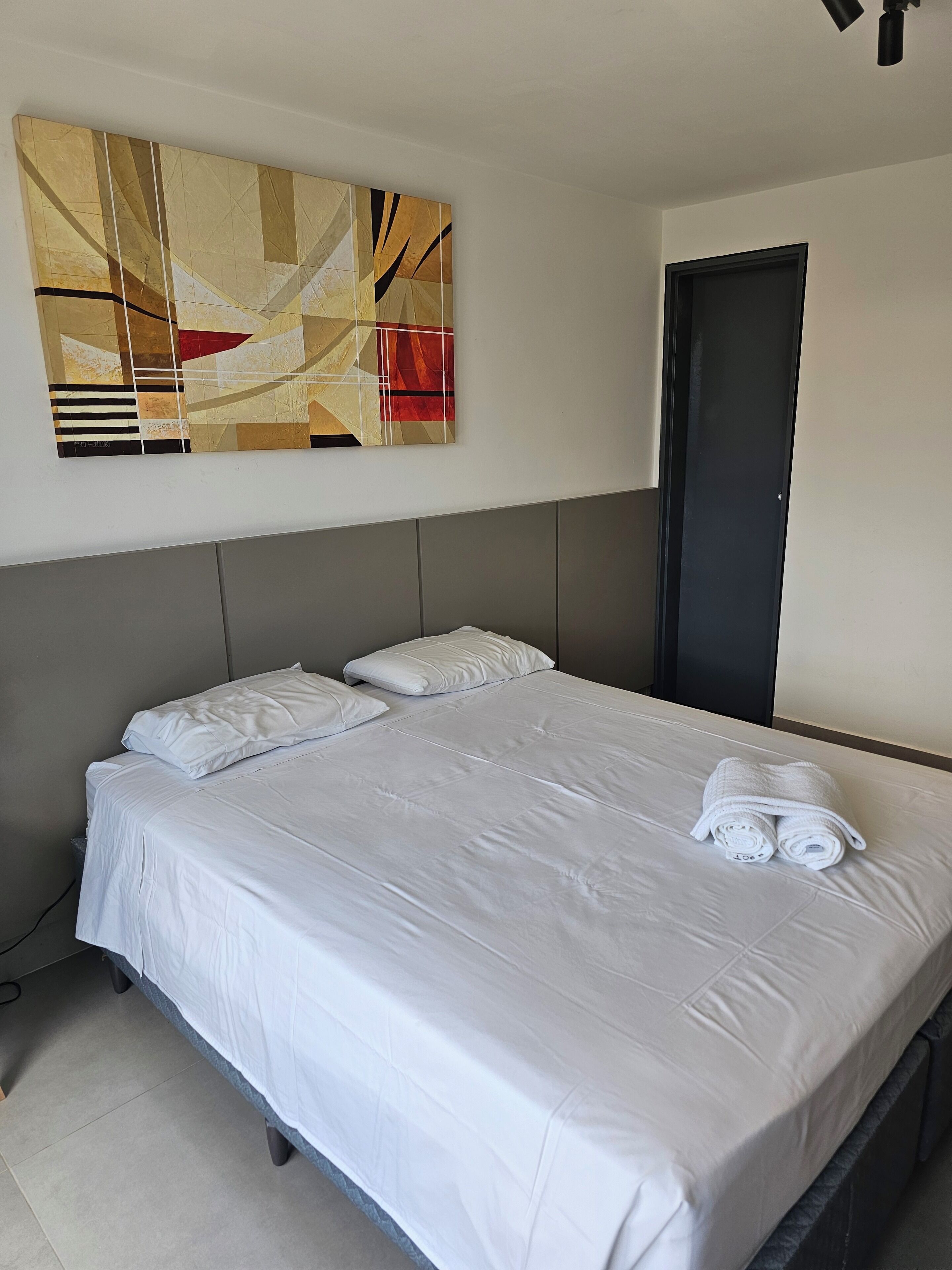 Una cama bien tendida con sábanas y almohadas blancas, un cuadro abstracto grande en la pared y una puerta cerrada.