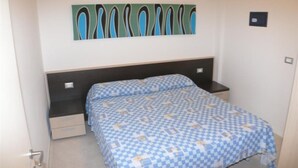Un dormitorio con cama, mesita de noche, cajón, una obra de arte en la pared y una puerta.