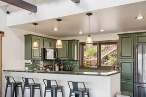 A kitchen with green cabinets, a stainless steel refrigerator, a large island with bar stools, pendant lighting, and a window with a view of the outdoors. - Views + Hot Tub, Firepit, Fireplace & PS5 (Banner Elk)
