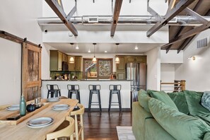 A modern kitchen with a central island, bar stools, and a green sofa in a spacious living area. - Views + Hot Tub, Firepit, Fireplace & PS5 (Banner Elk)