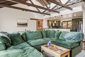 A green sectional sofa, a wooden coffee table with items, a kitchen with green cabinets, and a painting on the wall. - Views + Hot Tub, Firepit, Fireplace & PS5 (Banner Elk)