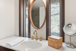 Ein Badezimmer mit weißem Waschbecken, goldenen Wasserhähnen, einer Holzausstattung, einem runden Spiegel und einem großen Fenster mit Blick auf Bäume.