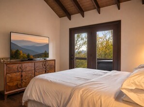 Ein Schlafzimmer mit Bett, Kommode, Fernseher und einer Tür zu einem Balkon mit Blick auf Bäume und Berge.