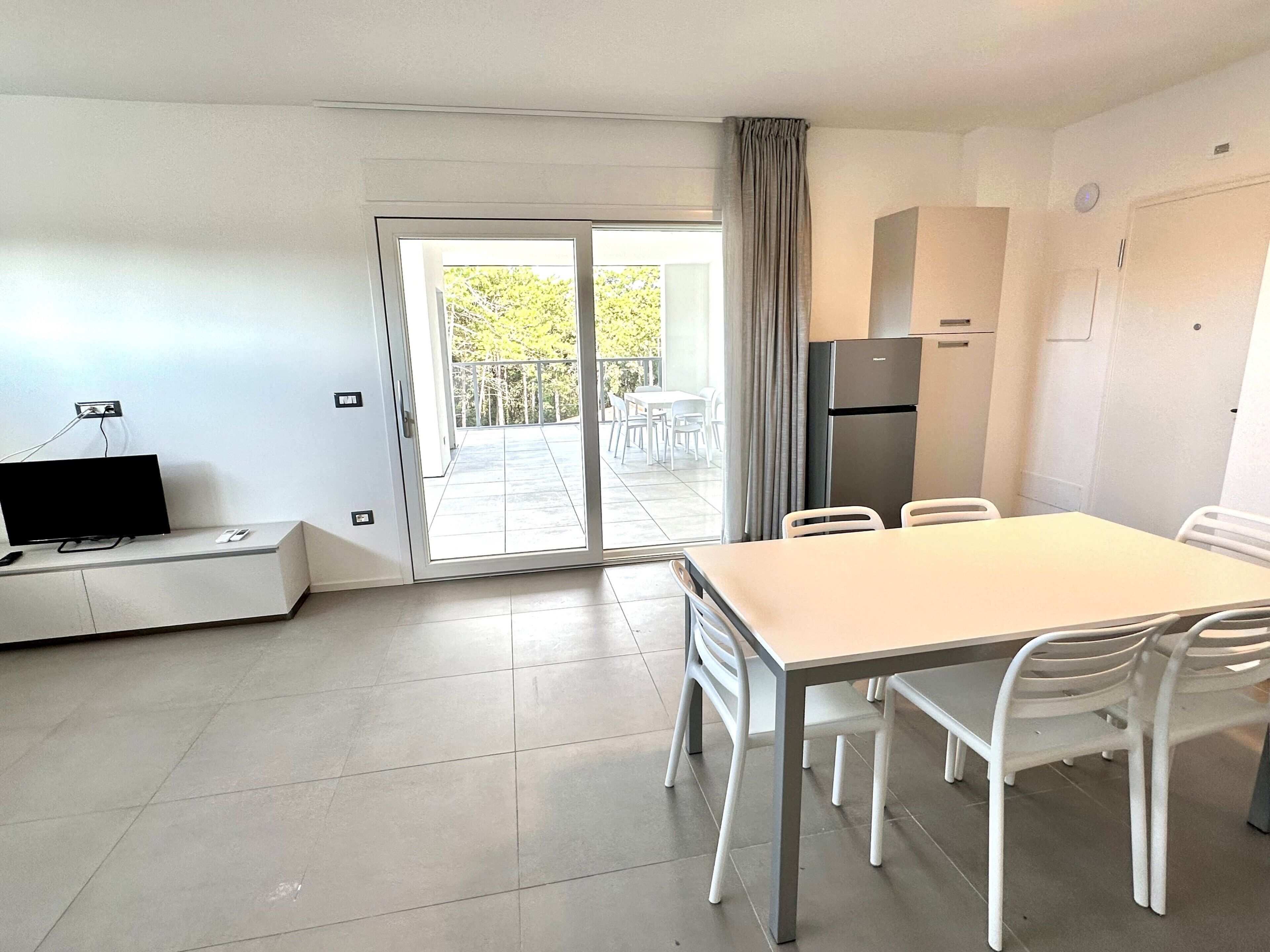 Een moderne keuken met een koelkast, een eettafel met stoelen, een televisie op een standaard en een schuifpui naar een buitenruimte met een tafel en stoelen.