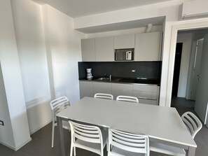 Eine moderne Küche mit weißen Schränken, einer Mikrowelle und einer Kaffeemaschine. Ein Esstisch mit vier Stühlen steht bereit.