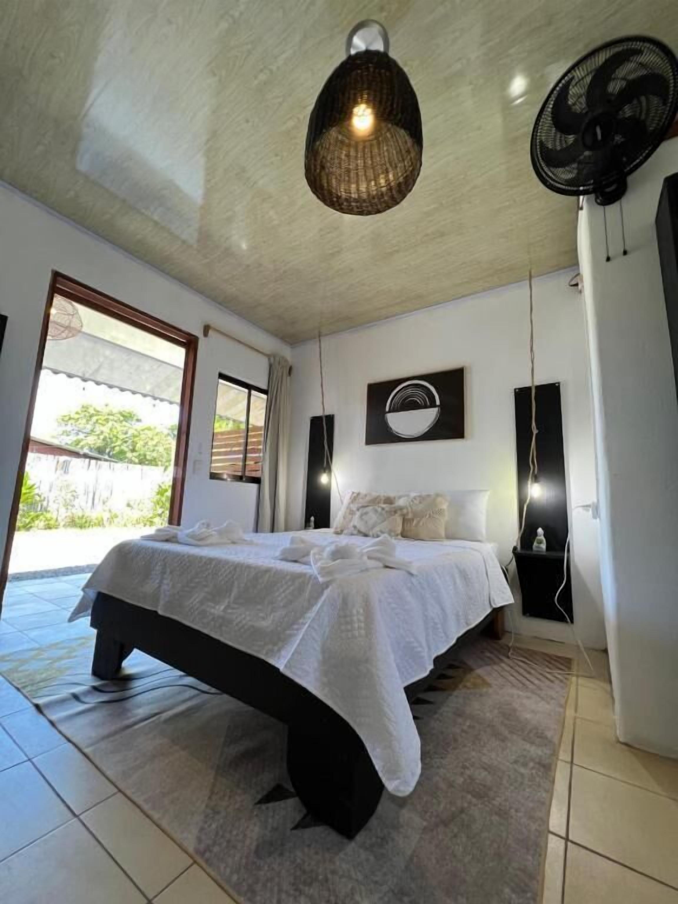 A bedroom with a bed, a ceiling fan, a wall-mounted light, and a view through a window.